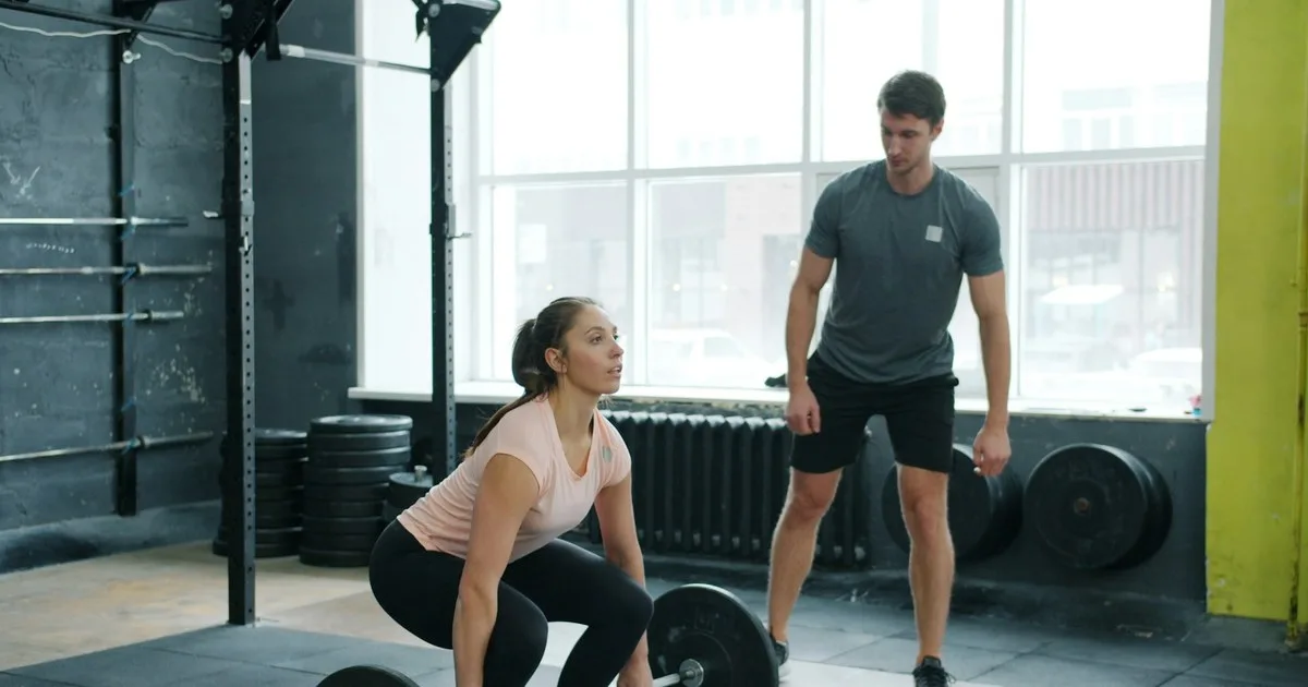 Una cliente prepara uno stacco da terra mentre il personal trainer, in piedi accanto a lei, ne supervisiona l'esecuzione.