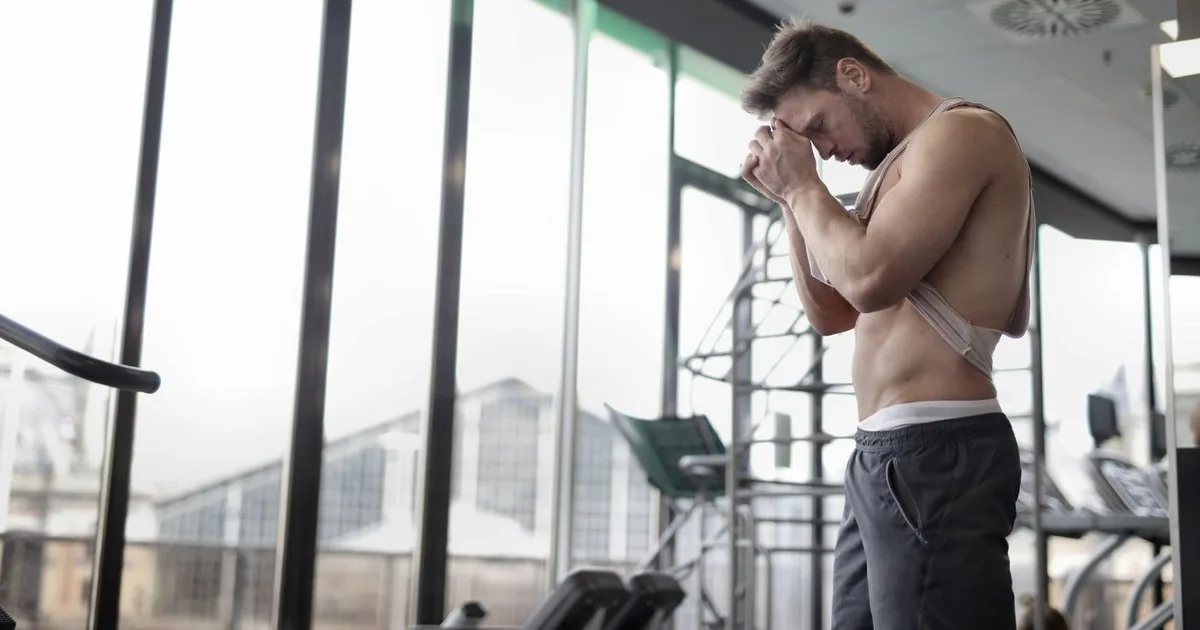 Un uomo con canottiera chiara sollevata, in piedi in una palestra con grandi vetrate, tiene le mani unite contro la fronte a testa china e occhi chiusi