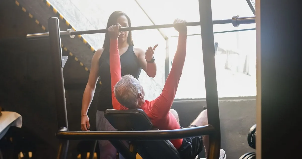 Un uomo anziano dai capelli bianchi, sdraiato su una panca in palestra, spinge verso l'alto un bilanciere mentre una trainer in piedi dietro di lui osserva l'esecuzione