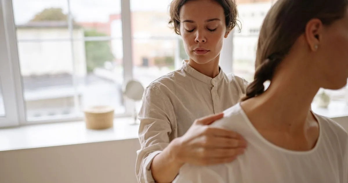 Una professionista in camicia beige appoggia una mano sulla spalla di una donna ripresa di spalle, in una stanza luminosa con una grande finestra