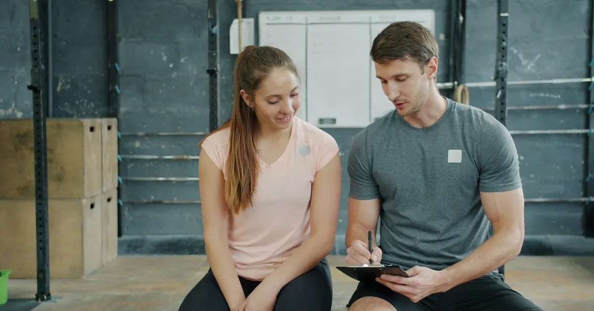 Un personal trainer prende appunti su un blocco durante il primo colloquio con una nuova cliente, seduti in palestra.