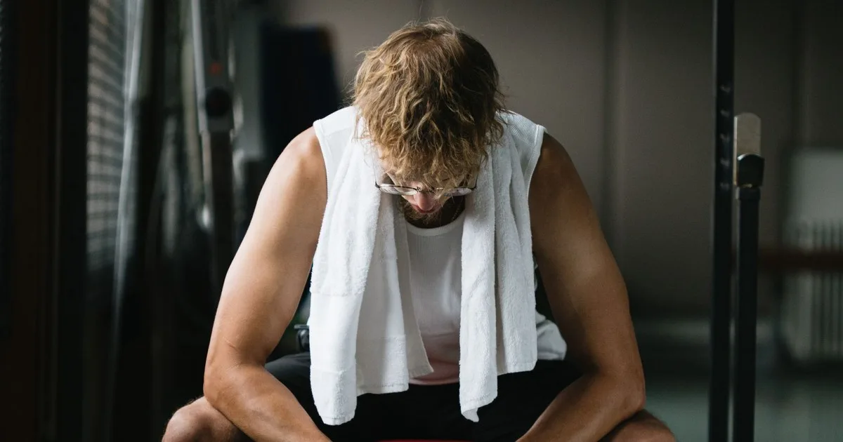 Uomo seduto a testa bassa su una panca in palestra, con un asciugamano bianco sulle spalle e un rack in secondo piano